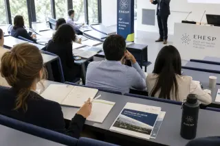 Group of adult students in a modern training amphitheater at a health school, seated with notebooks during a morning lecture, natural light and institutional colors creating a calm professional setting.