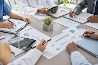 Directors and trainees collaborating over administrative documents in a bright, modern conference room of a healthcare facility, hands visible around a table during a budget planning session