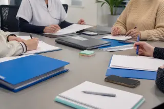 Morning meeting session in a neutral-toned conference room of a medico-social facility, professionals and trainees gathered around documents, maintaining anonymity and professionalism.