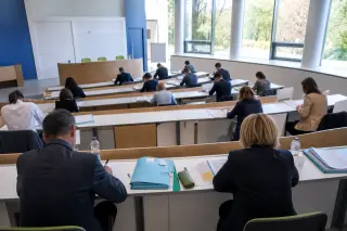 Morning exam session in a modern training amphitheater, candidates seated with folders, jury members observing, institutional colors, natural light, no visible faces.